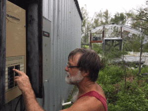 Photo of me wiring up new circuits for well, freezers, refrigerator, and chargers with tattered greenhouse in background.